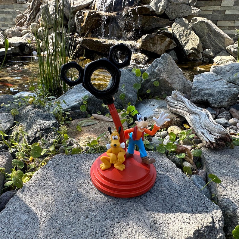 May include: A red and black Mickey Mouse-shaped desk organizer with a Goofy and Pluto figurine. The organizer has a base with three tiers and a Mickey Mouse head with ears on top. The figurine is of Goofy and Pluto standing on the base.