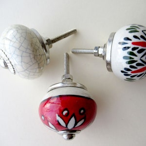 Puede incluir: Tres pomos de puerta de cerámica con diferentes diseños. Uno es blanco con un esmalte craquelado, otro es blanco con patrones geométricos rojos y verdes, y el último es blanco con un diseño floral rojo.