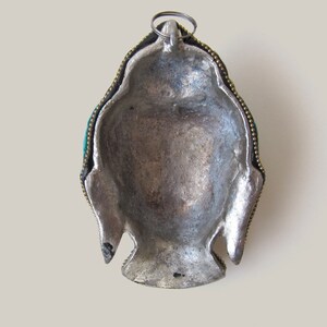 Op de afbeelding: Zilverkleurig metalen hanger met een Boeddha-gezichtsontwerp. De hanger heeft een groene steeninleg en goudkleurige accenten.