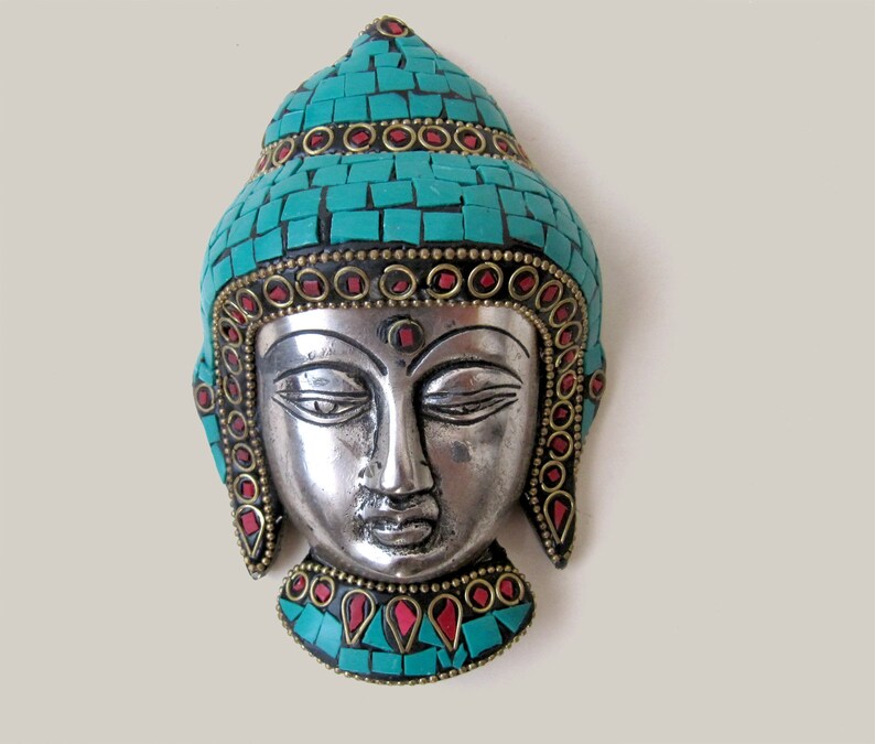 Op de afbeelding: Zilveren Boeddha-hoofd wanddecoratie met turquoise en rode moza&iuml;ekdetails. Het hoofd is omgeven door een decoratieve rand van turquoise en rode stenen.