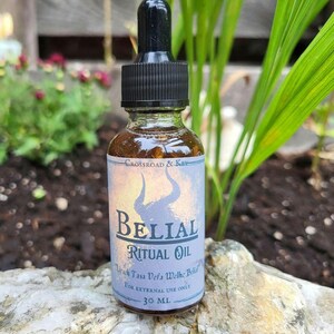 May include: A small, clear glass bottle with a black dropper top. The bottle is labelled "Belial Ritual Oil" with the text "Virach Tasa Vefa Wethe Belial" and "For External Use Only 30 ML".