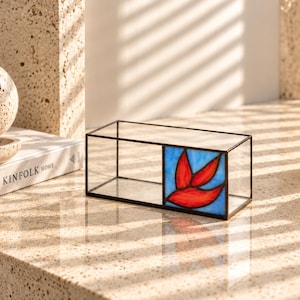 May include: A rectangular glass display box with a thin black metal frame. One side of the box features a stained glass panel with a vibrant red bird motif set against a light blue background, while the rest of the box is clear glass.