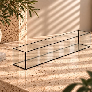 May include: A long, rectangular glass display box featuring a thin black metal frame. The structure is transparent with clear glass panels on all sides and an open top, designed as a sleek, elongated container for various small items.
