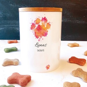 Könnte beinhalten: Weiße Keramik-Hundefutterdose mit Holzdeckel. Die Dose hat ein Aquarell-Pfotenabdruck-Design in Rosa-, Orange- und Gelbtönen. Der Text "Lunas Leckerli" ist auf die Dose gedruckt.