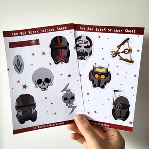 May include: Two sticker sheets featuring characters from the animated series "The Bad Batch". The sheets are white with red stars and feature various characters from the show, including a skull, a character with a red helmet, and a character with a red and black helmet. The text "The Bad Batch Sticker Sheet" is printed on each sheet.