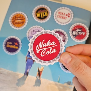 May include: A collection of nine round, colourful bottle cap stickers with various designs. One sticker features the text "Nuka Cola" in red lettering on a red background with a white border.