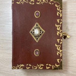 May include: A rectangular journal with a dark red cover featuring gold leaf-patterned borders and brass corner protectors. The center displays a white oval cabochon set in a gold diamond-shaped filigree frame, flanked by two small purple oval cabochons.