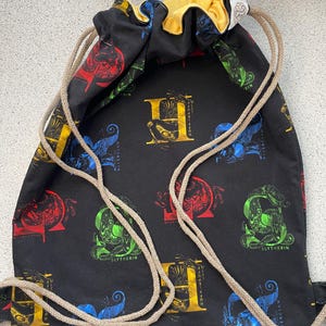 Pode incluir: Saco de cordão preto com forro amarelo. O saco é estampado com os brasões das casas de Hogwarts: Grifinória, Lufa-Lufa, Corvinal e Sonserina.
