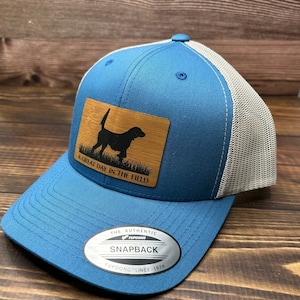 May include: A blue and gray baseball cap with a brown patch featuring a silhouette of a dog and the text "A Great Day In The Field". The cap has a silver label with the text "The Authentic Yupoong Snapback Yupoong Since 1974".