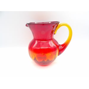 May include: A red glass pitcher with a yellow handle. The pitcher has a rounded body and a flared lip.