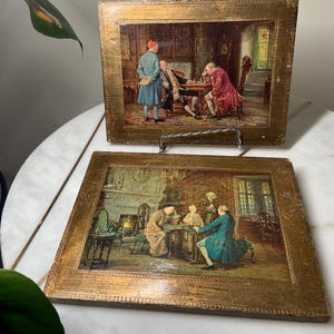 May include: Two framed vintage prints with gold-colored frames. The prints depict scenes of men in historical attire engaged in activities like playing chess. The artwork features detailed interior settings with rich colors and textures.