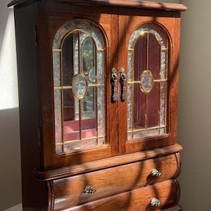 May include: A wooden jewelry armoire featuring two hinged doors with arched glass panels, gold-toned metal accents, and two lower drawers with small decorative knobs. The interior is lined with a solid red fabric, and the exterior has a polished finish.