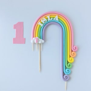 Pode incluir: Um topo de bolo de arco-íris com uma nuvem e o nome "Isla" em letras brancas. O arco-íris é feito de cores pastel e tem um redemoinho na parte inferior. Há um número "1" rosa em um palito separado.