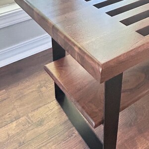 May include: A wooden bench with a dark brown finish and a lower shelf. The bench has a slatted top and is supported by black metal legs. The bench is placed on a hardwood floor.