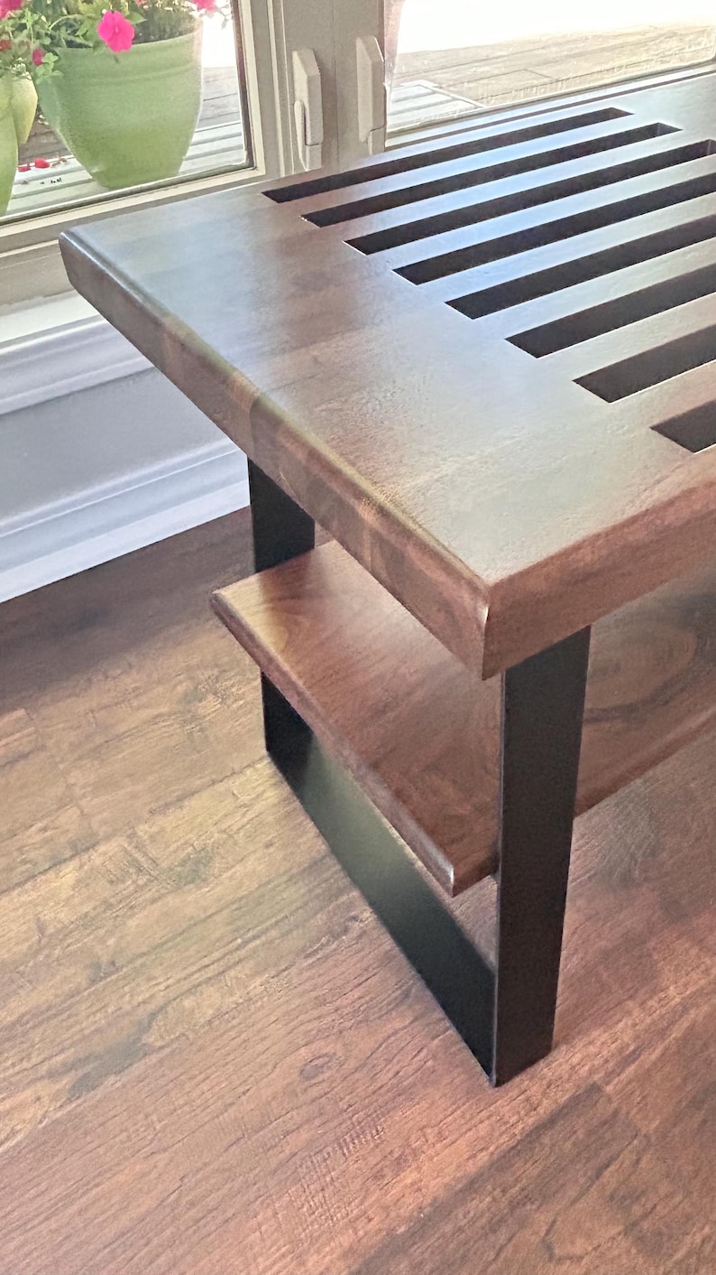 May include: A wooden bench with a dark brown finish and a lower shelf. The bench has a slatted top and is supported by black metal legs. The bench is placed on a hardwood floor.