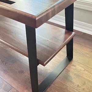 May include: A wooden bench with a dark brown finish and a slatted top. The bench has a lower shelf and is supported by black metal legs. The bench is placed on a hardwood floor.