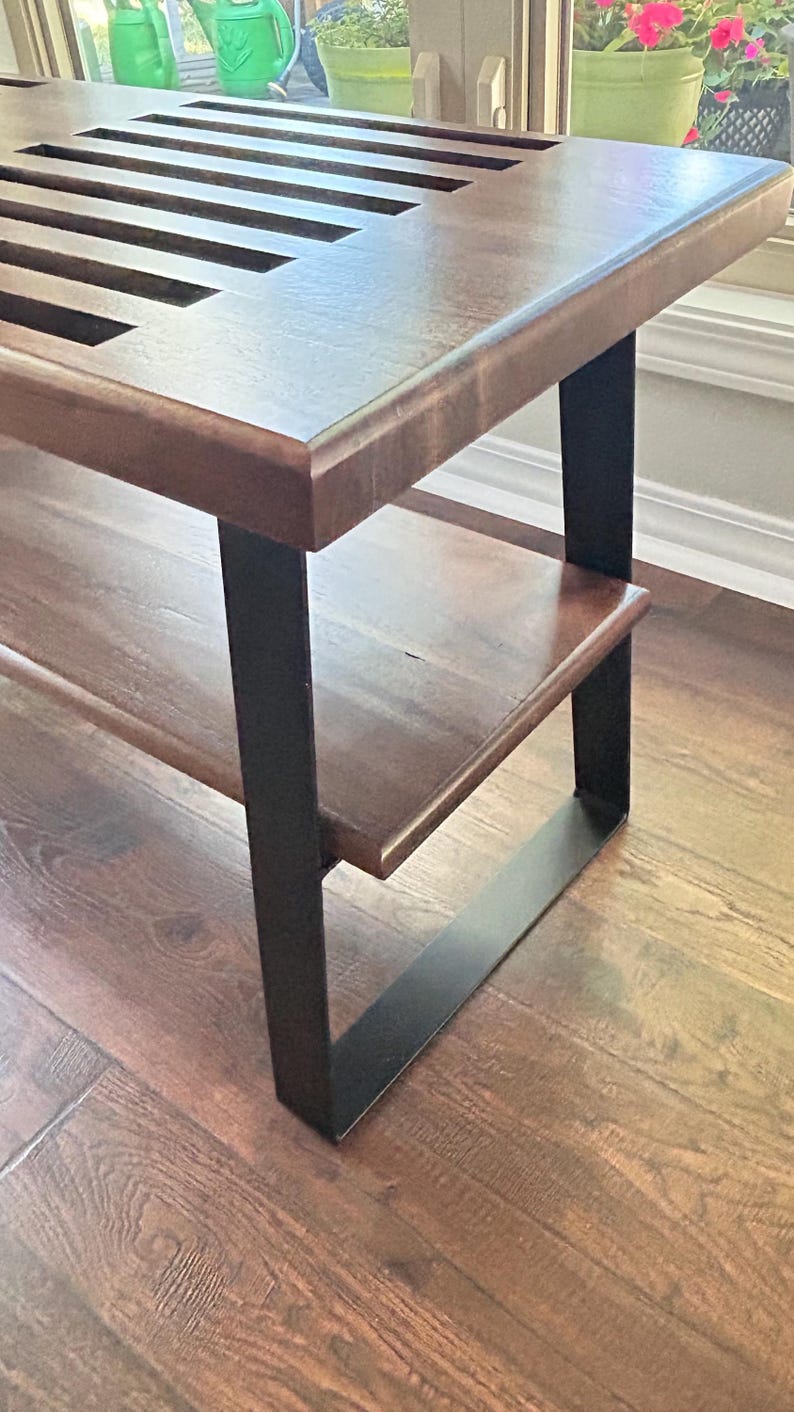 May include: A wooden bench with a dark brown finish and a slatted top. The bench has a lower shelf and is supported by black metal legs. The bench is placed on a hardwood floor.