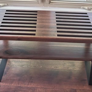 May include: A rectangular wooden coffee table with a dark brown finish. The tabletop features two sections of parallel cut-out slats. The table has a lower shelf and black metal legs. The table is approximately 48 inches long.