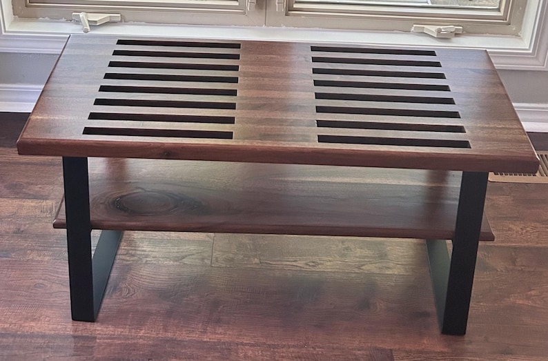 May include: A rectangular wooden coffee table with a dark brown finish. The tabletop features two sections of parallel cut-out slats. The table has a lower shelf and black metal legs. The table is approximately 48 inches long.