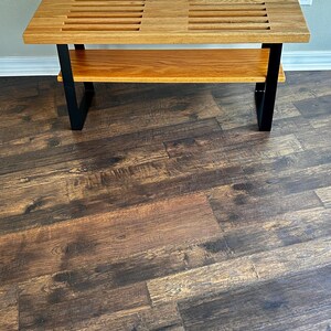 Puede incluir: Un banco de madera con una parte superior de listones y un estante inferior. El banco tiene patas de metal negro y est&aacute; sobre un suelo de madera.