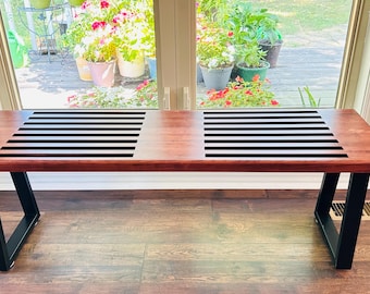 May include: A wooden bench with a slatted top and black metal legs. The bench is made of dark wood and has a natural finish. The bench is in front of a window with a view of a garden.