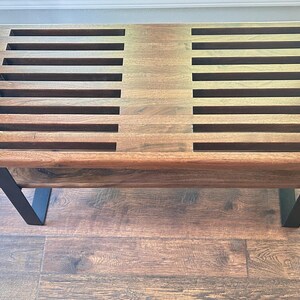 May include: A rectangular wooden coffee table with a dark brown finish. The tabletop features a series of evenly spaced rectangular cutouts. The table has black metal legs in a V-shape, providing support. The table is set on a hardwood floor.