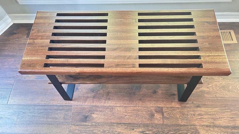 May include: A rectangular wooden coffee table with a dark brown finish. The tabletop features a series of evenly spaced rectangular cutouts. The table has black metal legs in a V-shape, providing support. The table is set on a hardwood floor.