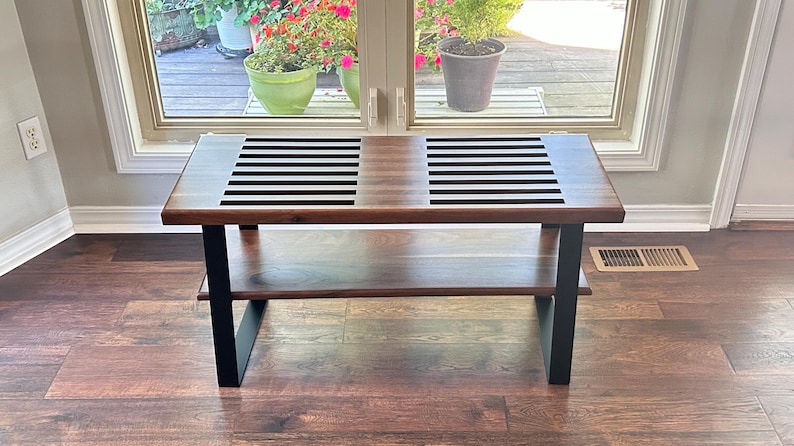 May include: A rectangular wooden coffee table with a lower shelf and black metal legs. The tabletop features a slatted design in the center. The table is set against a window with plants visible in the background.