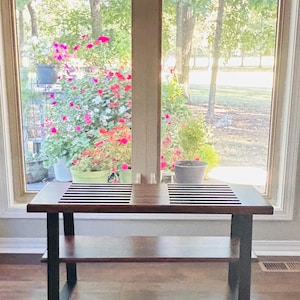 May include: A dark brown wooden coffee table with a lower shelf and black metal legs. The tabletop has a black and white striped design. The table is in front of a large window with a view of trees and flowers.