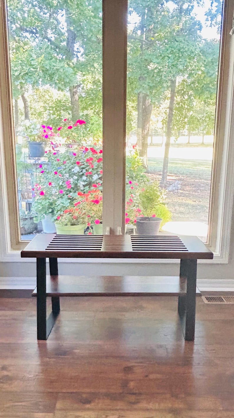 May include: A dark brown wooden coffee table with a lower shelf and black metal legs. The tabletop has a black and white striped design. The table is in front of a large window with a view of trees and flowers.