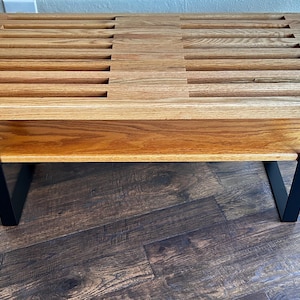 Puede incluir: Un banco de madera con una parte superior de listones y un estante inferior. El banco tiene patas de metal negro.