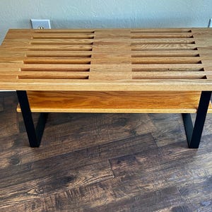 Puede incluir: Un banco de madera con una parte superior de listones y un estante inferior. El banco tiene patas de metal negro y est&aacute; hecho de madera clara.