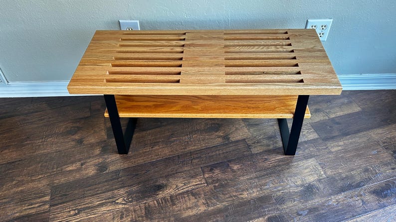 Puede incluir: Un banco de madera con una parte superior de listones y un estante inferior. El banco tiene patas de metal negro y est&aacute; hecho de madera clara.