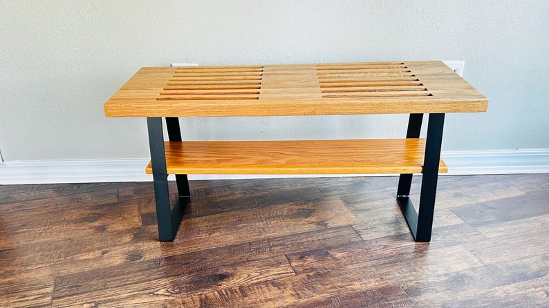 Puede incluir: Un banco de madera con una parte superior de listones y un estante inferior. El banco tiene patas de metal negro.