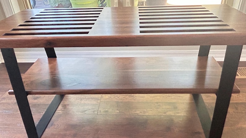 May include: A wooden coffee table with a dark brown finish. The tabletop features two sections with parallel slots. The table has a lower shelf and is supported by black metal legs. The table's dimensions are approximately 48 inches long, 24 inches wide, and 18 inches high.