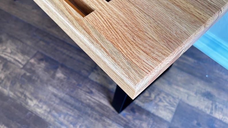 Puede incluir: Primer plano de una mesa de madera clara con acabado de veta de madera natural. La mesa tiene un borde biselado y est&aacute; sostenida por patas de metal negro.