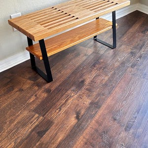 Puede incluir: Un banco de madera con un tablero de listones y un estante inferior. El banco tiene patas de metal negro y est&aacute; sobre un suelo de madera oscura.