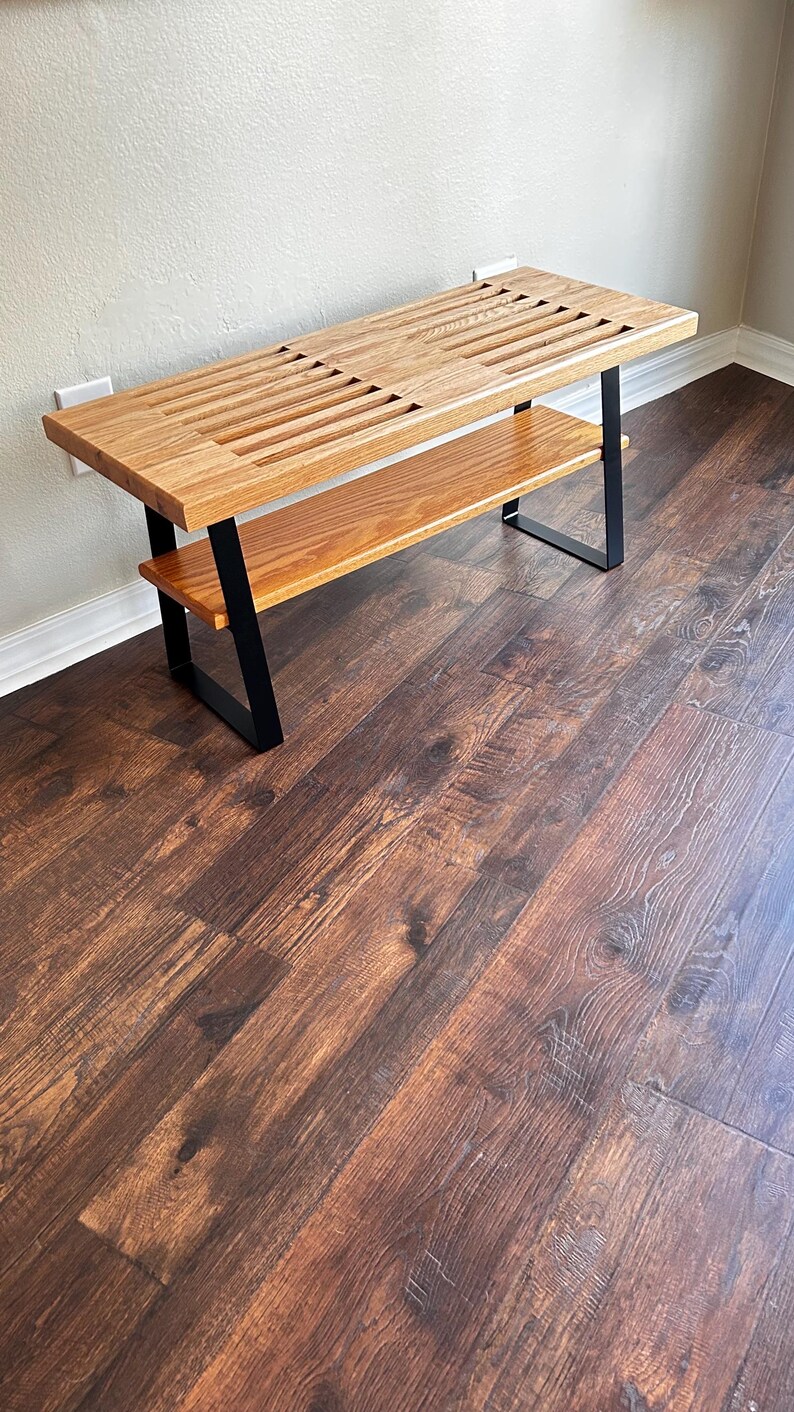 Puede incluir: Un banco de madera con un tablero de listones y un estante inferior. El banco tiene patas de metal negro y est&aacute; sobre un suelo de madera oscura.