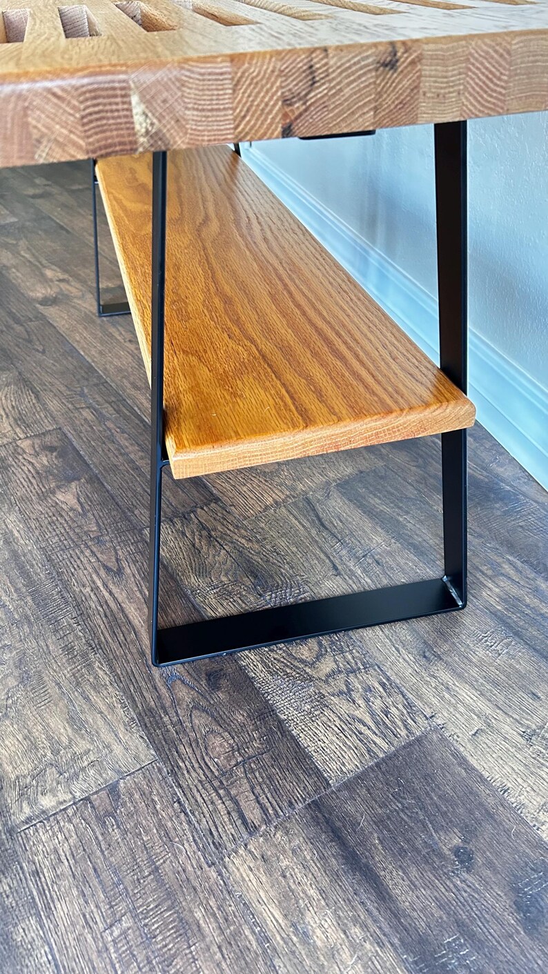 Puede incluir: Un banco de madera con una parte superior de listones y patas de metal negro. El banco tiene un estante inferior de madera.