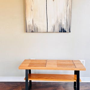 Puede incluir: Un banco de madera con un marco de metal negro y un estante inferior. El banco tiene una parte superior de listones y est&aacute; sentado en un suelo de madera.