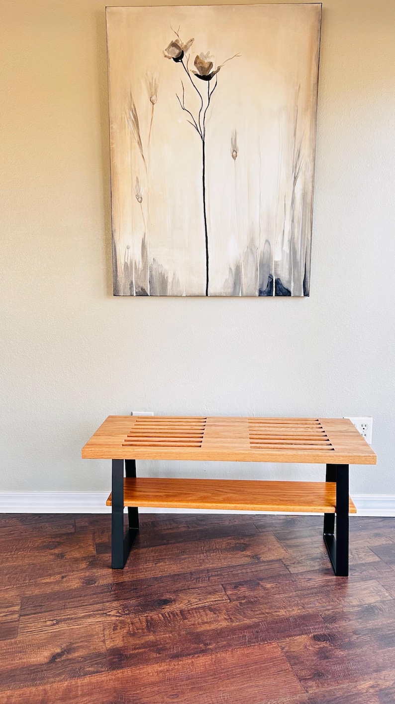 Puede incluir: Un banco de madera con un marco de metal negro y un estante inferior. El banco tiene una parte superior de listones y est&aacute; sentado en un suelo de madera.