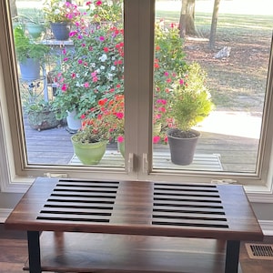 May include: A wooden bench with a dark brown finish and black metal legs. The bench has a slatted top and a lower shelf. The bench is placed in front of a window overlooking a garden with colorful flowers.