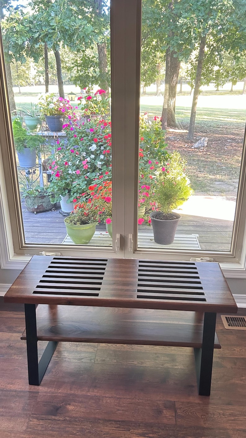 May include: A wooden bench with a dark brown finish and black metal legs. The bench has a slatted top and a lower shelf. The bench is placed in front of a window overlooking a garden with colorful flowers.