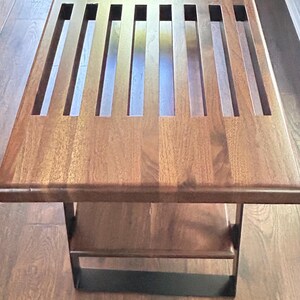 May include: A rectangular wooden coffee table with a dark brown finish. The tabletop features a series of evenly spaced rectangular cutouts. The table has a lower shelf and a black metal base. The table is approximately 48 inches long.