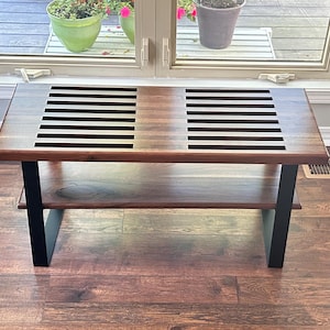 May include: A rectangular wooden coffee table with a lower shelf. The tabletop features two sections with black horizontal slats. The legs are black, and the table is set on a hardwood floor. The table is approximately 48 inches long.