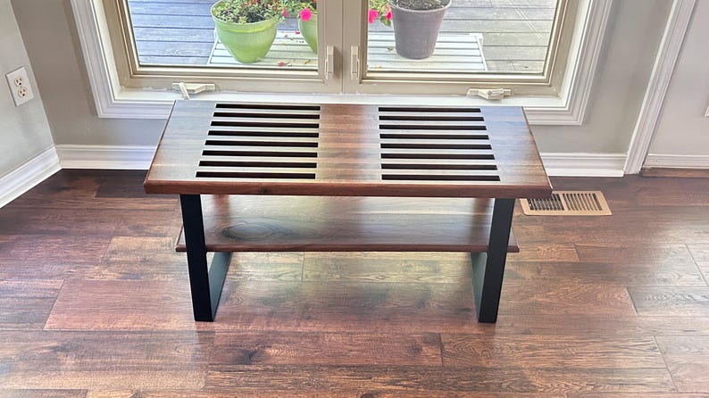 May include: A rectangular wooden coffee table with a lower shelf. The tabletop features two sections with black horizontal slats. The legs are black, and the table is set on a hardwood floor. The table is approximately 48 inches long.