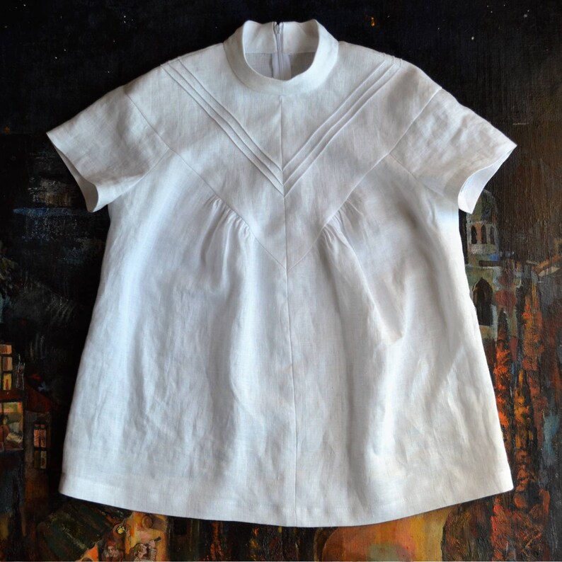 May include: A white linen blouse with a high neckline and short sleeves. The blouse has a gathered front and a centre seam.