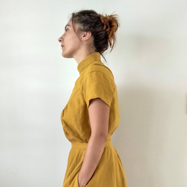May include: A woman wearing a mustard yellow dress with a high neck and short sleeves. The dress has a gathered skirt and a pocket on the left side.