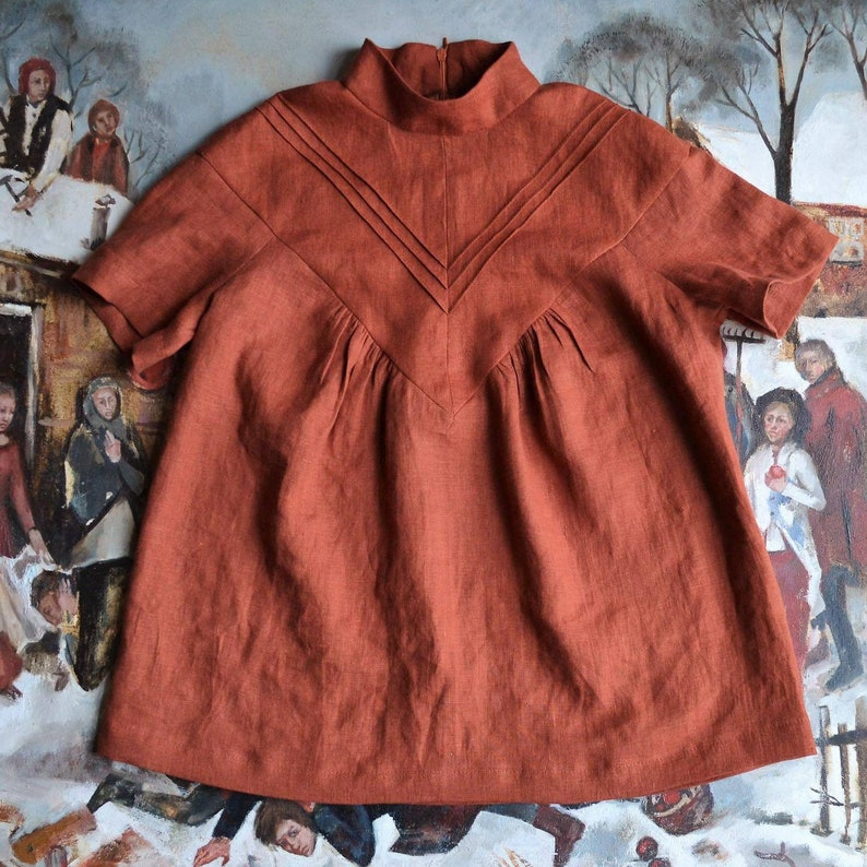 May include: A rust-coloured linen blouse with a high neckline and short sleeves. The blouse has a V-shaped neckline with pleats and a decorative seam detail.