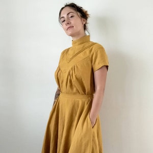 May include: A mustard yellow linen dress with a high neck and short sleeves. The dress has a gathered bodice and a full skirt with pockets.